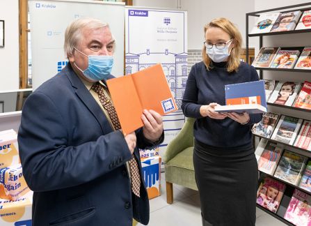 A photograph. The director of the villa Decius institute for culture, Dominika Kasprowicz, gives the books to the director of the Krakow library, Stanisław Dziedzic. In the backgound a tall shelf full of books.
